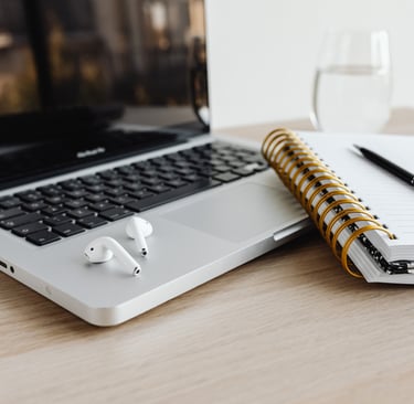 Open laptop with air pods on a desk with notebooks, pen, and glass of water. 