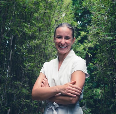 Portrait d'Anaïs plein sourire en blouse de soin beige dans une allée de bambous