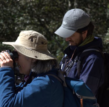 A birdwatcher wearing a hat looks through binoculars while Chiapas birding guide Valente takes notes