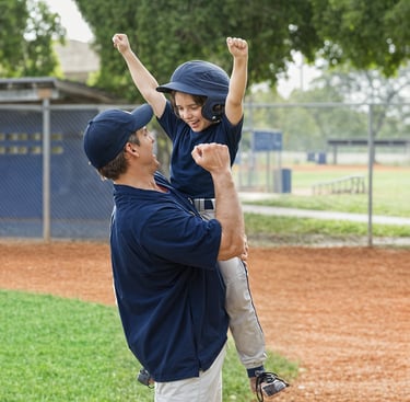 A parent-coach lifts a his player to celebrate on the baseball field.