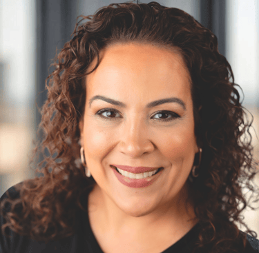 Professional headshot of a smiling woman with curly brown hair and gold hoop earrings.