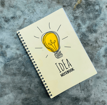 Un carnet à spirale beige avec une illustration d'ampoule jaune et le titre 'Idea Notebook'.