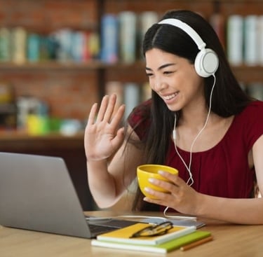 Female student learning French online, wearing headset, smiling and waving at her laptop