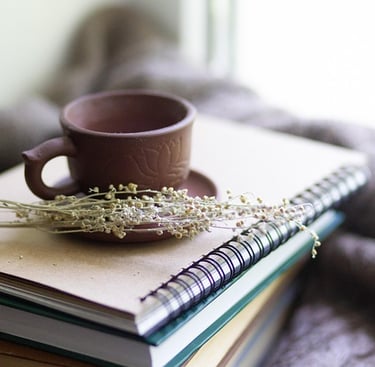 Rustic clay teacup on a stack of spiral notebooks with dried flowers and a cozy blanket.