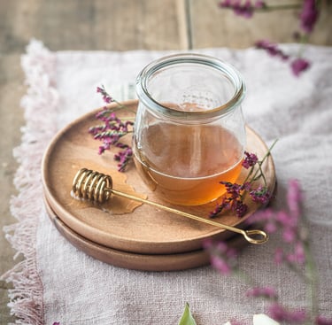 Golden organic honey in a glass jar with a metal dipper and purple flowers on a rustic wooden table.