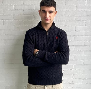 a man in a sweater and jeans standing in front of a white brick wall
