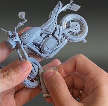 Hands sanding a 3D printed grey resin miniature motorcycle model to remove support marks.