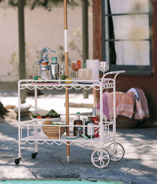 A white vintage-style bar cart with summer cocktails and snacks by a swimming pool under a green striped umbrella.