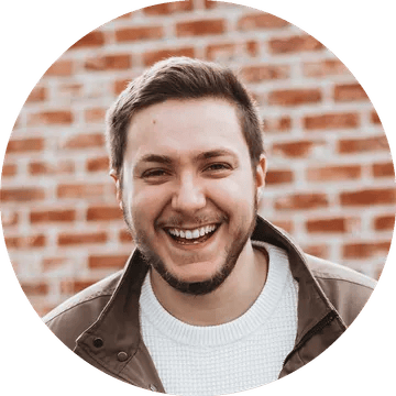 Smiling white man with short brown hair and facial stubble wearing a brown jacket and white shirt in front of a brick wall.