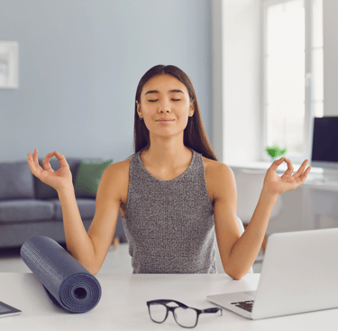 A female professional meditating in front her desk at home, featuring a computer and a yoga mat.