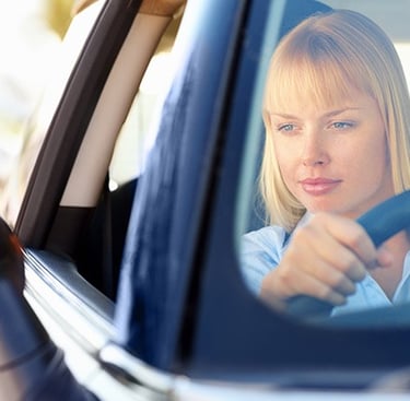 A blonde woman driving a car and looking ahead while holding the steering wheel on a sunny day.