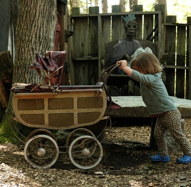 A toddler pushes a vintage stroller in a spooky forest setting near a skeletal king prop.