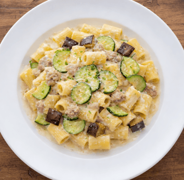 Rigatoni pasta with zucchini, eggplant, and sausage in a creamy sauce on a white plate.