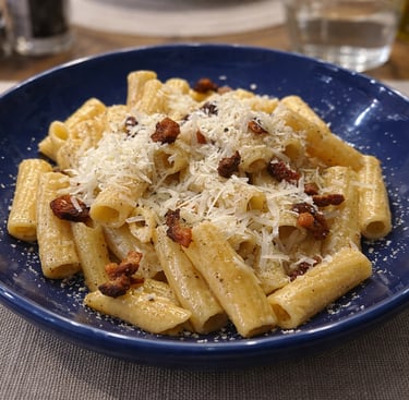 Rigatoni pasta alla gricia topped with pecorino cheese and crispy guanciale in a blue bowl.