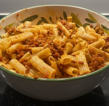 A ceramic bowl filled with rigatoni pasta tossed in a hearty meat bolognese sauce on a black marble surface.