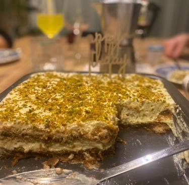 Homemade pistachio tiramisu cake topped with crushed nuts and a happy birthday sign on a dining table.