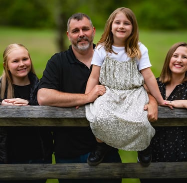A happy family of four poses for a portrait outdoors behind a rustic wooden fence in a lush green field.
