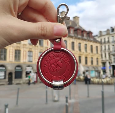 Red leather Vagabond Life travel keychain with engraved rings held in a city square.
