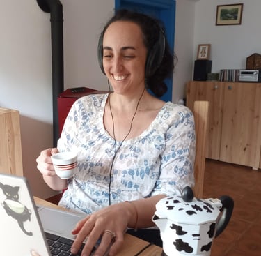 Smiling woman wearing headphones while working on a laptop and enjoying a cup of coffee at home.