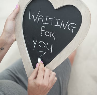 Pregnant woman writing Waiting For You on a heart-shaped chalkboard for a maternity photo shoot.
