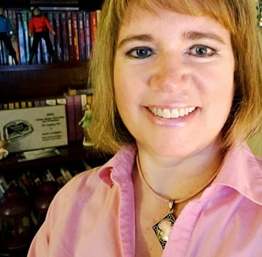 Susan Gable wearing a pink shirt, smiling, posing in front of her bookshelf.