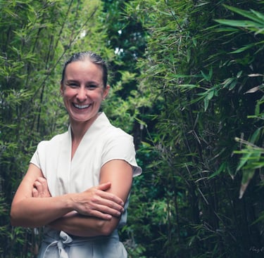 Portrait d'Anaïs plein sourire en blouse de soin beige dans une allée de bambous
