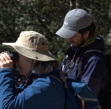 A birdwatcher wearing a hat looks through binoculars while Chiapas birding guide Valente takes notes