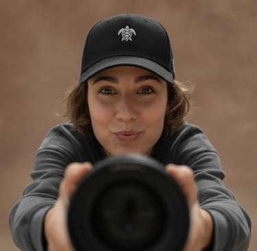 A smiling woman wearing a black sea turtle logo baseball hat holds a professional camera lens toward the viewer.