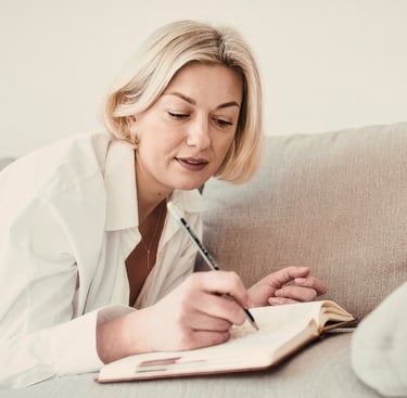 A blonde woman in a white shirt lying on a couch while writing in a journal or notebook.