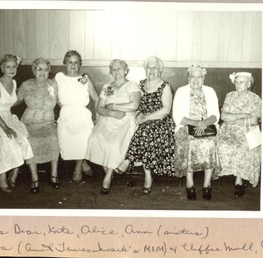 Vintage black and white photo of seven women in floral dresses sitting together at a formal gathering.