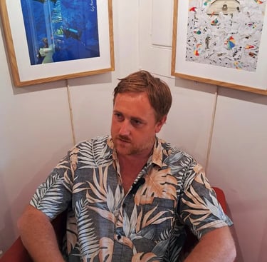 Artist seated in a red chair beneath framed digital prints of underwater and courthouse-themed artwo