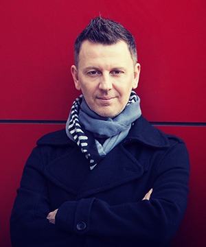 Portrait of a John Taylor in a navy pea coat and scarf against a vibrant red wall.