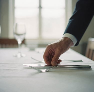 Close-up of a hand in a dark suit adjusting polished silver on an immaculate dining table setting, soft natural light, shallow depth of field, editorial film photography, 35mm