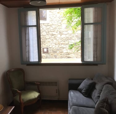 Rustic living room with a grey sofa, green armchair, and open window overlooking Quillan Church.