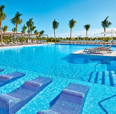 Luxury tropical resort swimming pool with submerged lounge chairs, palm trees, and sunny blue sky.