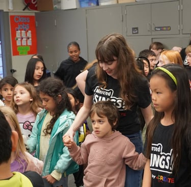 Diverse elementary students and teacher gathered in a classroom for a Merced Sings music program activity.