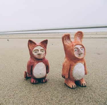 two small ceramic animals sitting on a sandy beach