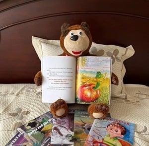 A smiling teddy bear holding an open picture book with four other books on the bedspread.
