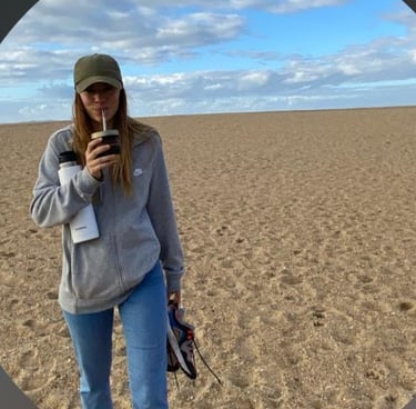 a woman in a hat and sunglasses is drinking mate and walking on the beach