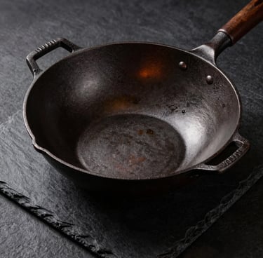 High-contrast editorial shot of an uncoated cast iron wok on a dark slate surface, showing raw iron texture, dramatic side lighting
