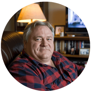 A middle-aged man in a red plaid shirt sitting in a leather chair in a cozy home library.