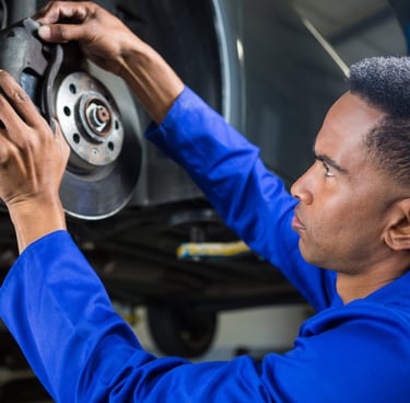 A professional auto mechanic in blue coveralls inspecting a car's brake disc and caliper in a repair shop.