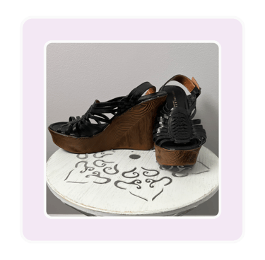 Pair of black strappy leather wedge sandals with high wood platform heels on a white stand.