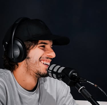 aledima sorride mentre parla al microfono durante un podcast, con cuffie e cappello nero