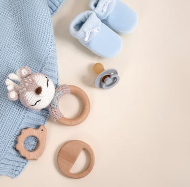 Flat lay of blue baby booties, a knit blanket, pacifier, and wooden teething toys on a beige background.