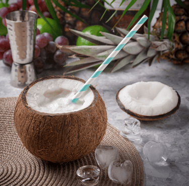 A tropical coconut drink with a striped straw, served in a coconut shell with ice cubes and pineapple nearby.
