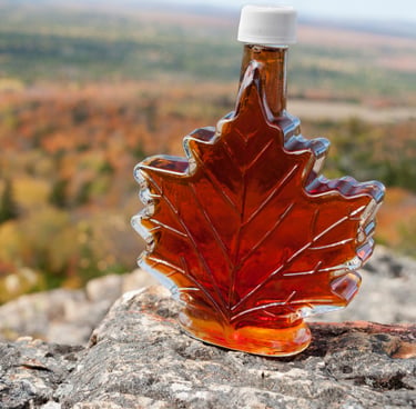 A maple leaf-shaped glass bottle of amber maple syrup resting on a stone with blurred autumn trees behind.