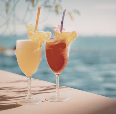 Two tropical cocktails (one yellow, one red) with straws and pineapple slices by the hazy blue ocean.