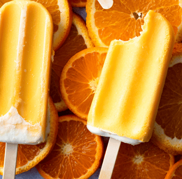 Two orange cream popsicles, one with a bite taken out, resting on a background of fresh, juicy orange slices.