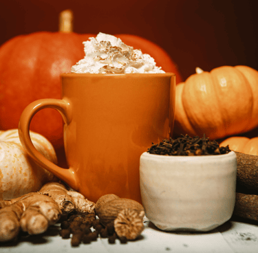 An orange mug of hot pumpkin spice drink topped with whipped cream, surrounded by whole pumpkins, ginger, and spices.
